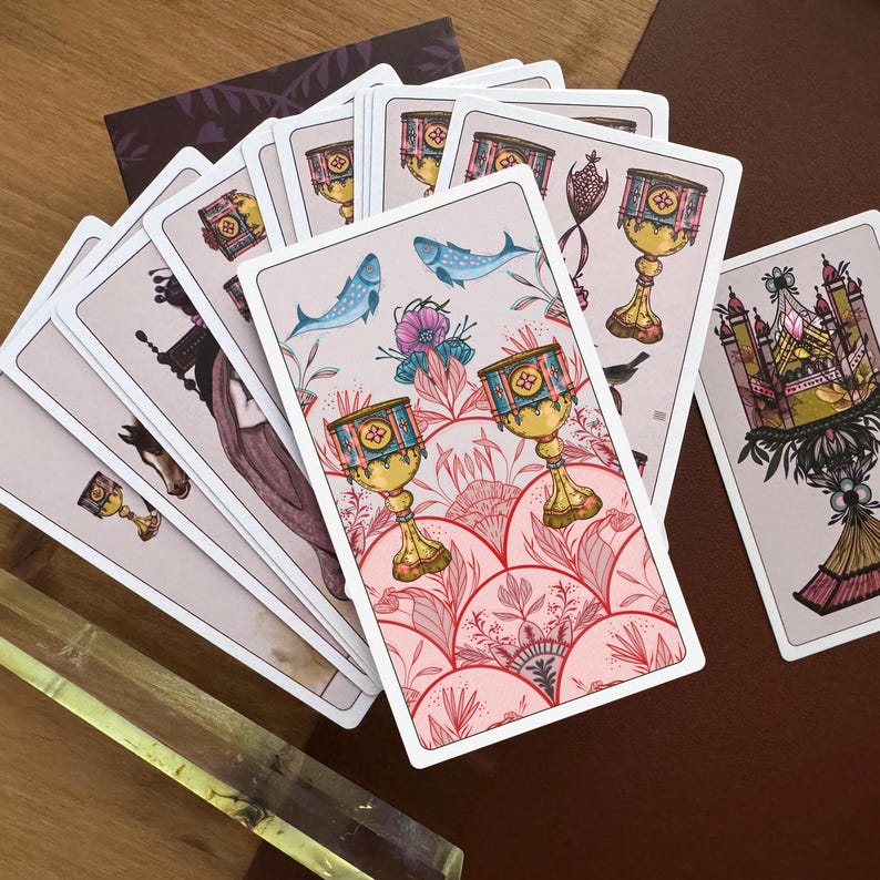 May include: A deck of tarot cards fanned out, featuring a central card with two golden chalices, blue fish, and pink floral patterns. The cards display intricate illustrations of cups, architectural structures, and figures in a soft colour palette.