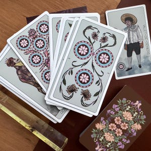 May include: A collection of tarot cards featuring light grey backgrounds with intricate floral and circular patterns in pink and blue. One card displays a figure in a straw hat labelled Valet de Deniers, while another shows a floral back design.
