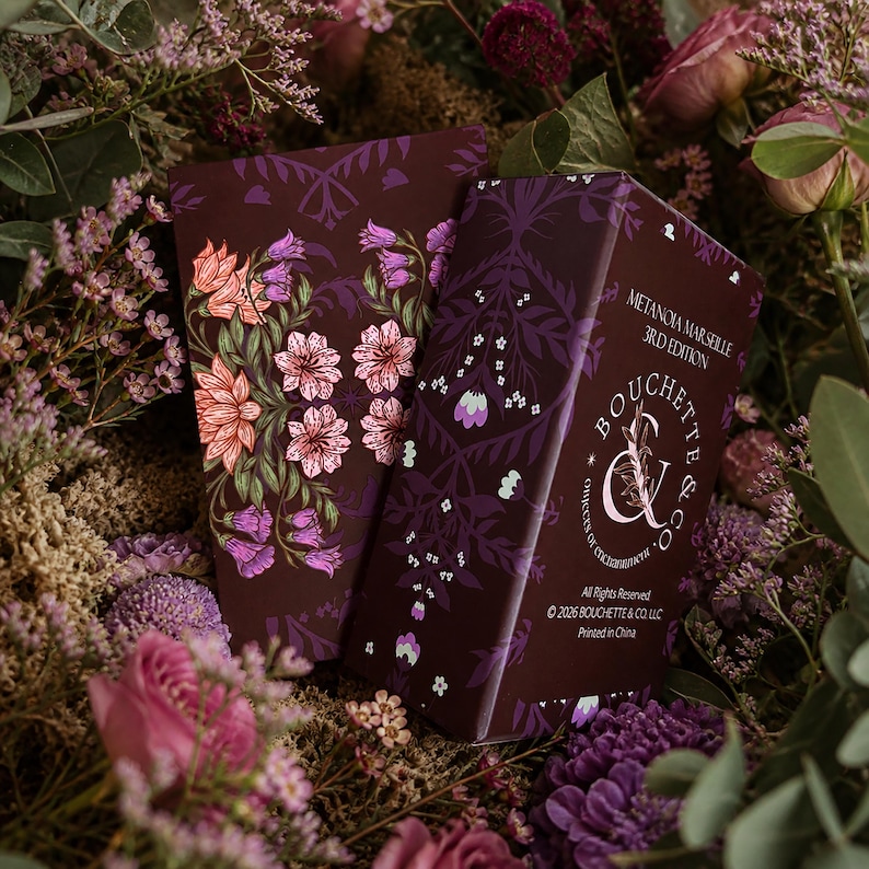 May include: A rectangular deck of cards box in deep purple with a floral pattern of pink and purple flowers. The box features the text Metanoia Marseille 3rd Edition, Bouchette & Co., and copyright information printed in white on the front surface.
