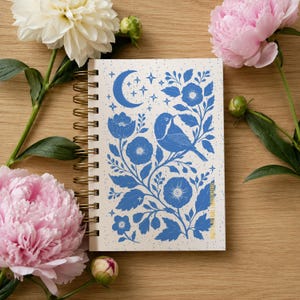 May include: A spiral-bound notebook with a blue floral and bird design. The cover features a crescent moon, stars, and botanical illustrations. The notebook is surrounded by pink and white peonies on a wooden surface.
