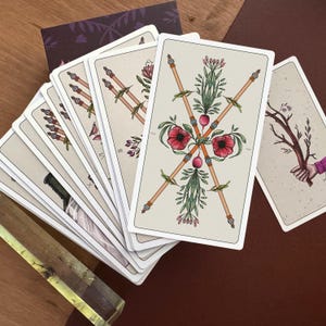 May include: A deck of tarot cards fanned out, featuring a prominent card with two crossed wooden wands adorned with red poppy flowers and green foliage. The cards have a cream-coloured background with detailed botanical illustrations and thin borders.