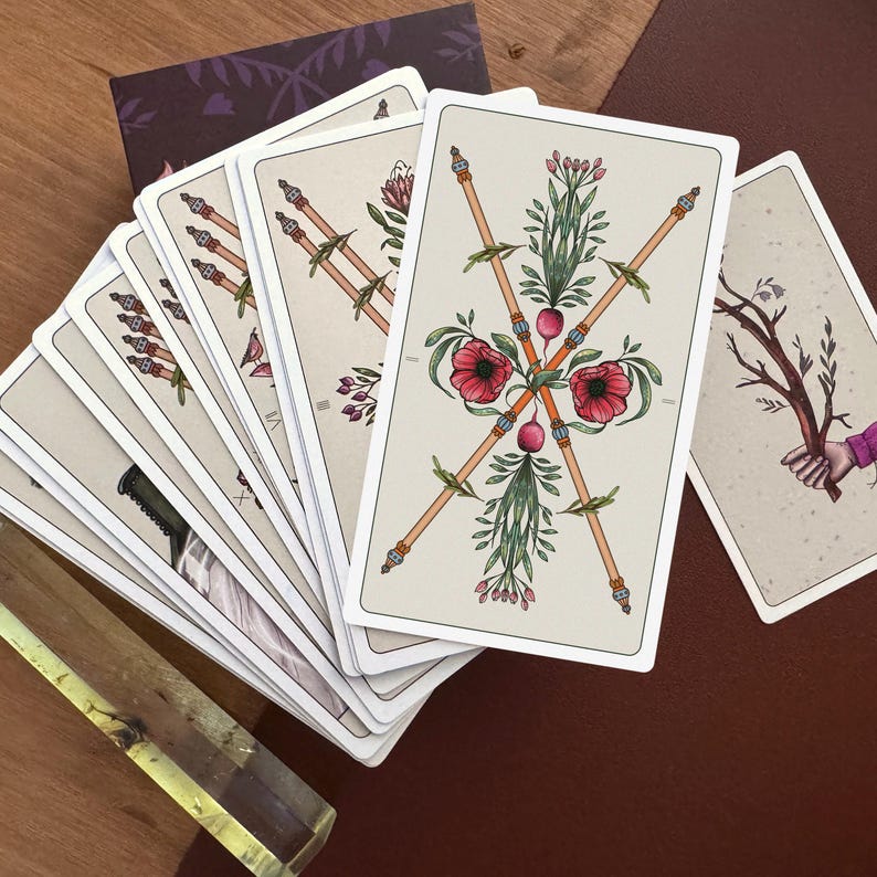 May include: A deck of tarot cards fanned out, featuring a prominent card with two crossed wooden wands adorned with red poppy flowers and green foliage. The cards have a cream-coloured background with detailed botanical illustrations and thin borders.