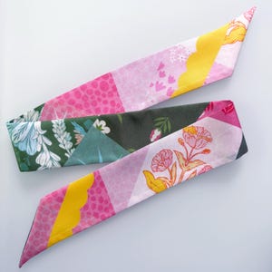May include: A long, narrow fabric hair scarf featuring a patchwork design of various patterns. The print includes sections of dark green with white floral motifs, bright pink with polka dots, yellow scalloped shapes, and light pink with flowers.