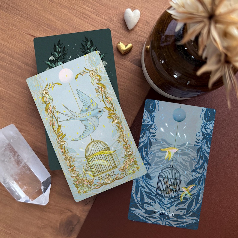 May include: Two rectangular tarot cards featuring illustrations of birds and cages. The left card is light blue with a bird flying above a cage and the text "RELEASE". The right card is dark blue with a bird inside a cage and the text "RESTRAIN".