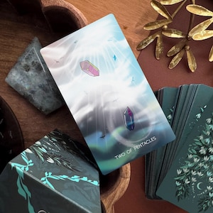 May include: A Two of Pentacles tarot card featuring a light blue sky with two floating crystals, one pink and one dark blue, surrounded by small birds. The card has white text at the bottom and is part of a deck with dark green floral backs.