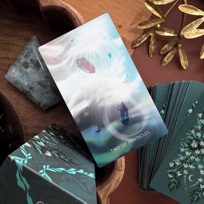 May include: A Two of Pentacles tarot card featuring a light blue sky with two floating crystals, one pink and one dark blue, surrounded by small birds. The card has white text at the bottom and is part of a deck with dark green floral backs.