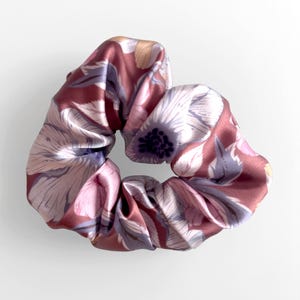 May include: A floral scrunchie in shades of burgundy, pink, and lavender. The fabric features a repeating pattern of stylized flowers and leaves. The scrunchie is made of a soft, silky material, perfect for adding a touch of elegance to any hairstyle.