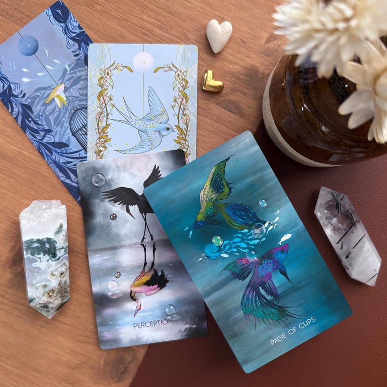 May include: A collection of tarot cards featuring artistic illustrations of a bird, a crane, and a betta fish, with the text Perception and Page of Cups. The cards are arranged alongside two crystal points, a small white heart, and a gold heart.