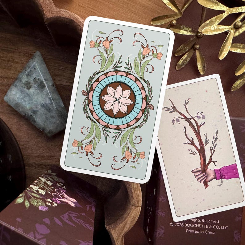May include: Two rectangular tarot cards with rounded corners. One card features a light green background with a central circular mandala design in pink, blue, and brown, surrounded by floral vines. The other card shows a hand holding a brown branch.