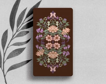 May include: A rectangular card with a dark brown background featuring a symmetrical, intricate botanical illustration. The design includes light pink lotus flowers, purple bell-shaped blossoms, green leaves, and thistle-like elements.