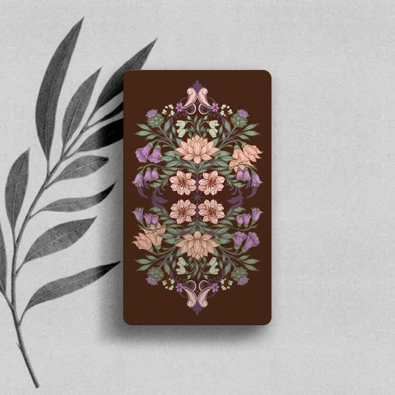 May include: A rectangular card with a dark brown background featuring a symmetrical, intricate botanical illustration. The design includes light pink lotus flowers, purple bell-shaped blossoms, green leaves, and thistle-like elements.