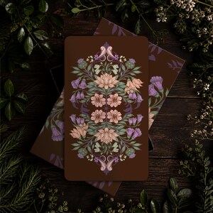 May include: A rectangular card with a dark brown background featuring a symmetrical, intricate botanical illustration. The design includes light pink lotus-like flowers, purple bell-shaped blossoms, and green foliage arranged in a vertical pattern.