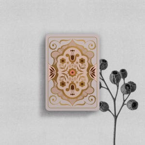 May include: A rectangular playing card featuring a symmetrical floral pattern in shades of brown, gold, and muted pink. The design includes stylized flowers, leaves, and swirling lines centered on a light mauve background with rounded corners.