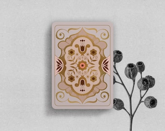 May include: A rectangular playing card featuring a symmetrical floral pattern in shades of brown, gold, and muted pink. The design includes stylised flowers, leaves, and swirling lines centred on a light mauve background with rounded corners.