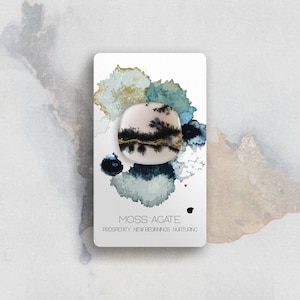 May include: A smooth, rounded moss agate stone featuring dendritic black inclusions resembling tree branches on a cream-colored base. The stone is mounted on a white display card with watercolor-style blue and gold accents and the text MOSS AGATE PROSPERITY NEW BEGINNINGS NURTURING.