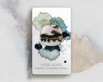 May include: A smooth, rounded moss agate stone featuring dendritic black inclusions resembling tree branches on a cream-coloured base. The stone is mounted on a white display card with watercolour-style blue and gold accents and the text MOSS AGATE PROSPERITY NEW BEGINNINGS NURTURING.