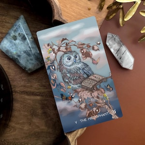 May include: A rectangular tarot card titled V THE HIEROPHANT featuring a detailed illustration of a grey owl perched on a branch with an open book, butterflies, and small yellow flowers against a soft blue and cloudy sky background.