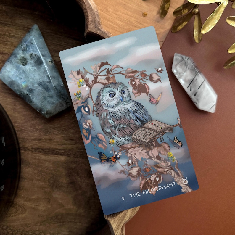 May include: A rectangular tarot card titled V THE HIEROPHANT featuring a detailed illustration of a grey owl perched on a branch with an open book, butterflies, and small yellow flowers against a soft blue and cloudy sky background.