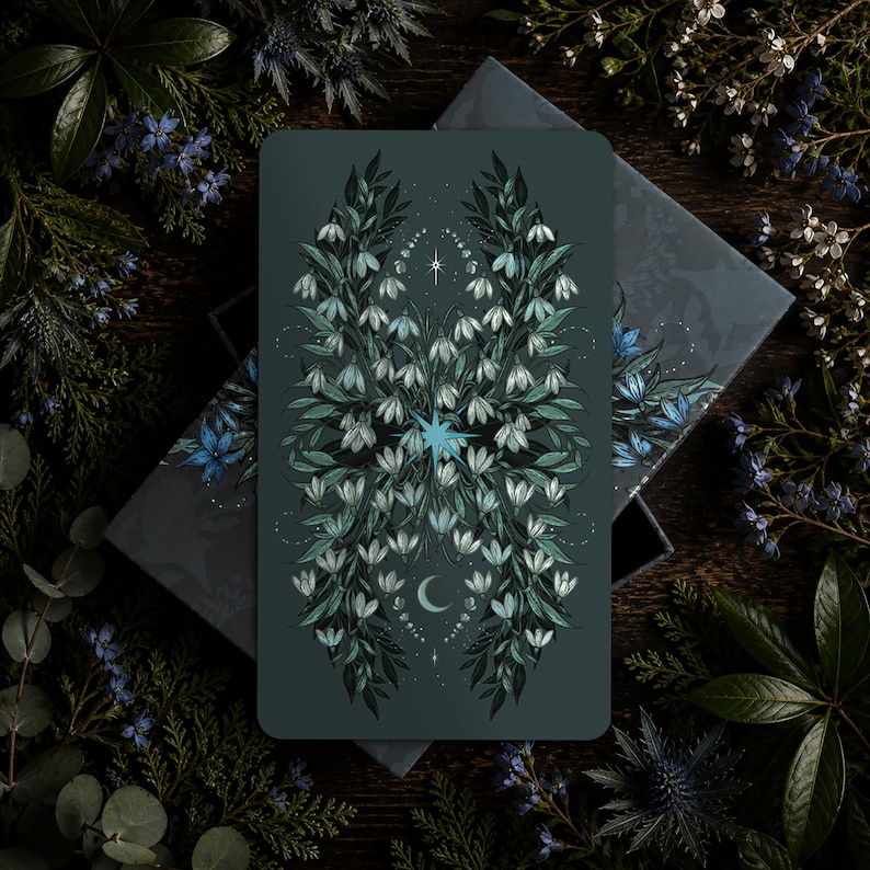 May include: A rectangular tarot card with a dark teal background featuring a symmetrical illustration of white snowdrop flowers, green leaves, a central blue starburst, a small crescent moon, and delicate white star accents on the surface.