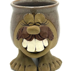 May include: A whimsical ceramic mug with a face design. The mug features a brown and tan color scheme, with large white teeth, a brown mustache, and feet. The mug is designed to look like a smiling face.