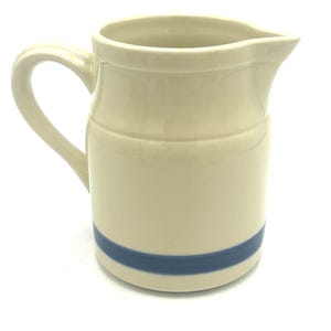 May include: Cream-colored ceramic pitcher with a curved handle and spout. A horizontal blue stripe encircles the base. The pitcher has a classic, simple design, suitable for serving beverages.