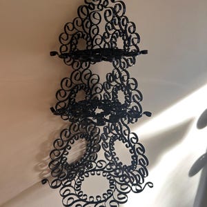 May include: Black wall-mounted shelf with a decorative, swirling design. The shelf has three tiers and is made of a dark material. The design features numerous loops and curls, creating an ornate, artistic appearance. The shelf is mounted on a light-colored wall.