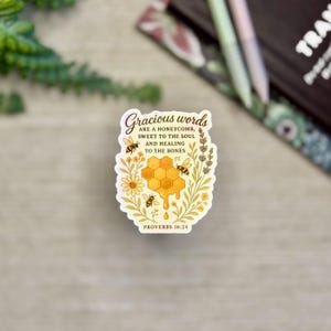 May include: A sticker with the text "Gracious words are a honeycomb, sweet to the soul and healing to the bones" and "Proverbs 16:24". The sticker features a honeycomb design with bees and floral accents in yellow, brown, and green.