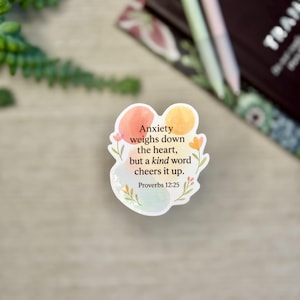 May include: A sticker with the text "Anxiety weighs down the heart, but a kind word cheers it up. Proverbs 12:25." The sticker has a white background with watercolor-style orange, yellow, and blue shapes and floral accents.
