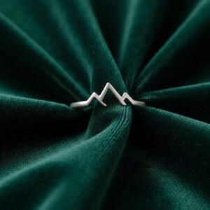 May include: A silver ring with a mountain range design. The ring is set against a dark green velvet background. The mountain range is a simple, stylized outline.