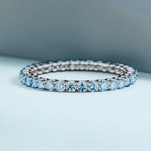 Op de afbeelding: Een zilveren ring met een doorlopende band van lichtblauwe edelstenen. De ring wordt getoond tegen een zachte blauwe achtergrond, die de glans van de stenen benadrukt. De edelstenen zijn gelijkmatig verdeeld over de gehele omtrek.