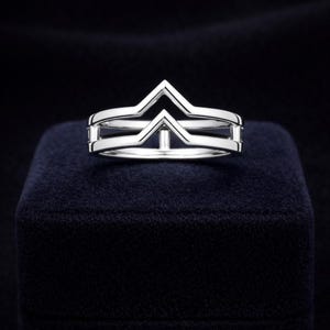 Könnte beinhalten: Ein silberner Ring mit geometrischem Design, mit einer zentralen V-Form und zwei parallelen Bändern. Der Ring wird auf einer dunkelblauen Samtoberfläche präsentiert, wodurch ein Kontrast entsteht, der seinen metallischen Glanz hervorhebt. Dieser Schmuck eignet sich für den Alltag.