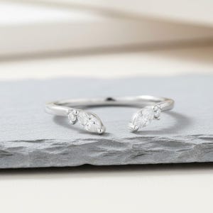 Peut inclure: Une bague en argent au design ouvert, ornée de deux diamants taille marquise et de petites pierres d'accent. La bague est posée sur une surface grise, mettant en valeur son style délicat et élégant. Un bijou parfait pour toutes les occasions.
