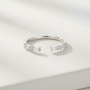 Peut inclure: Une bague ouverte argentée ornée d'une rangée de pierres précieuses rectangulaires et rondes. La bague présente un design ouvert, avec un espace entre les deux extrémités. Les pierres sont serties sur le dessus de la bague.