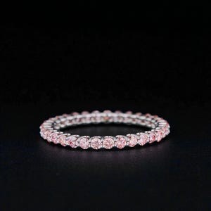 Puede incluir: Un delicado anillo con una fila continua de piedras preciosas rosas engastadas en una banda plateada. El anillo se muestra sobre un fondo negro, resaltando el brillo de las piedras preciosas. Una pieza de joyería fina.