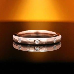 May include: Rose gold band ring with five small, round, clear gemstones evenly spaced around the band. The ring is highly polished and reflects the warm, golden light in the background. The ring is sitting on a reflective surface.