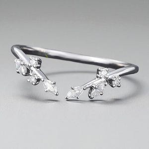 Könnte beinhalten: Ein silberfarbener Ring mit einem einzigartigen, offenen Design. Der Ring hat zwei dekorative, blattähnliche Zweige, die jeweils mit mehreren klaren, funkelnden Edelsteinen besetzt sind. Die Edelsteine sind eine Mischung aus runden und marquiseförmigen Schliffen, was die Eleganz des Rings unterstreicht.