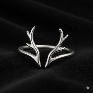 Puede incluir: Un anillo plateado con un diseño estilizado de cuernos de ciervo. Los cuernos del anillo están abiertos en la parte delantera, creando un aspecto único y elegante. El anillo se encuentra sobre un fondo oscuro, resaltando su brillo metálico.