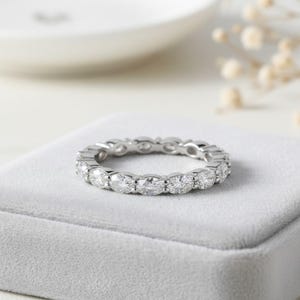 以下が含まれることがあります： オーバルカットのダイヤモンドがセットされたシルバーのエタニティリング。リングはグレーのベルベットの表面に置かれ、背景には白い皿と小さな白い花がぼやけています。ダイヤモンドは透明で輝いています。