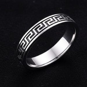 Può includere: Un anello d'argento con un motivo a chiave greca attorno alla fascia. L'anello è molto lucido e riflette la luce, su uno sfondo scuro. Il design è un motivo geometrico continuo e ripetitivo.