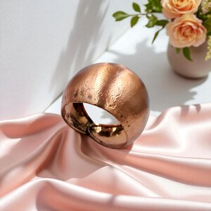Pode incluir: Uma pulseira de metal texturizado cor de cobre, com formato arredondado e abobadado. A pulseira está sobre um tecido de cetim rosa, com um vaso de rosas cor de pêssego ao fundo. A pulseira tem um acabamento em metal martelado.