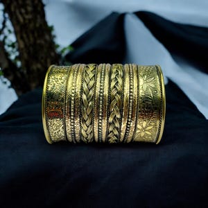 Peut inclure: Un bracelet manchette doré avec des motifs complexes. Le bracelet présente des motifs floraux et tressés, créant un aspect texturé. Le métal a une finition antique, lui donnant un aspect vintage. Le bracelet est présenté sur une surface noire.