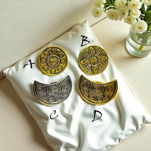 May include: Four decorative metal pieces with intricate patterns, labelled A, B, C, and D. Two are circular, and two are crescent-shaped. The pieces are gold and silver, resting on a white cushion. A vase of white flowers is in the background.
