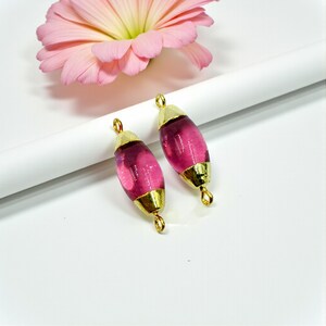 May include: Two pink glass beads with gold-coloured caps and loops. The beads are oval-shaped and have a translucent quality. A pink flower is in the background. These beads are suitable for jewellery making.