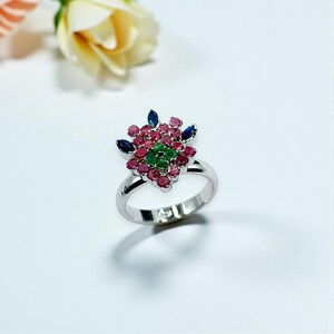 May include: A silver ring with a floral design. The ring has a cluster of small, round, red gemstones, with green gemstones at the centre. Blue gemstones are placed around the red gemstones. The ring is set against a white background.
