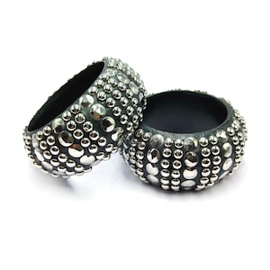 May include: Two dark gray, studded bangles. The bangles are round with a black interior and are covered in silver-colored, round studs. The bangles are displayed on a white background.