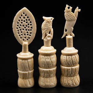 May include: Three off-white carved objects with cylindrical bases featuring textured, grooved patterns. The left object has a flat, oval, lattice-cut top. The centre object features a perched owl carving. The right object features a standing lion carving.