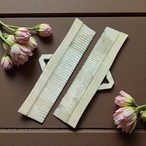 May include: Two off-white combs with a handle are displayed on a brown surface, alongside pink flowers. The combs have fine teeth and a decorative design. The combs are approximately 7 inches long.