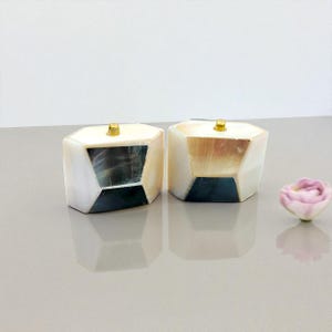 May include: Two decorative, geometric boxes with a pearlescent finish and brass-colored knobs. Each box has a dark, angular inlay. A small, pink flower is in the background. The boxes are on a reflective surface.