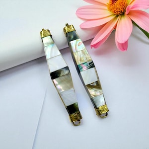 May include: Two elongated, decorative jewelry pieces with a mosaic design. The pieces are made of rectangular mother-of-pearl tiles in shades of white, black, and iridescent colors. They have gold-colored metal caps and loops. A pink flower is in the background.