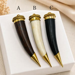 May include: Three horn-shaped pendants labelled A, B, and C. Pendant A is dark brown, B is off-white, and C is black. Each pendant features a gold-toned metal cap with a beaded texture at the top and a pointed gold-toned metal tip at the bottom.
